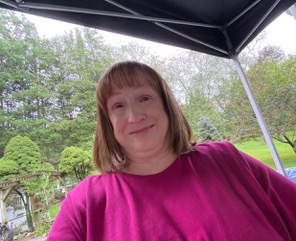 Jenine is a white, middle age woman with shoulder-length reddish brown hair wearing a dark pink shirt. She is smiling. There are trees and green vegetation behind her.
